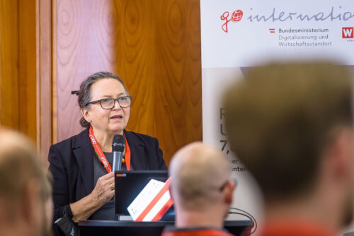 In the foreground, three heads of the audience are out of focus from behind; in front of them, Karin Stieldorf speaks into a microphone.