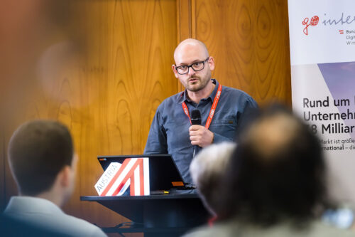 In the foreground, three heads of the audience are out of focus from behind; in front of them, Christoph Treberspurg speaks into a microphone.