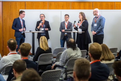 audience seen from behind sitting in front of 5 people who are standing at two tables, one person,Karin Stieldorf is talking into a microphone.