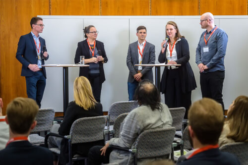 audience seen from behind sitting in front of 5 people who are standing at two tables, one person, Astrid Schneider is talking into a microphone.